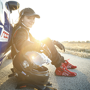 Natalie Fenaroli, wearing her racing gear, sits next to a car as the sun sets.