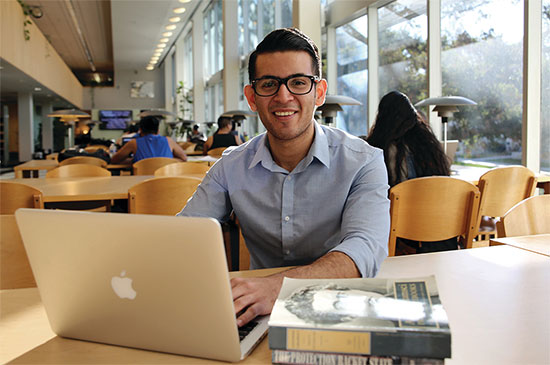 Roberto Bonilla sits at his laptop in Wardman Library, with two stacked books nearby.