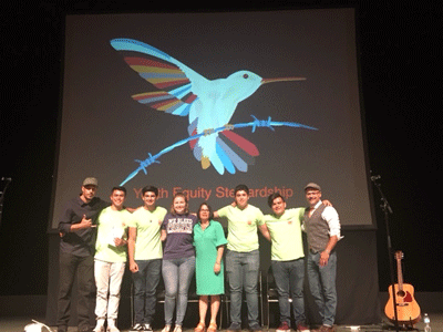 a group of 8 people stand on a stage in front of a screen. on the screen is a projected image of a bird with the words "youth faculty stewardship" underneath