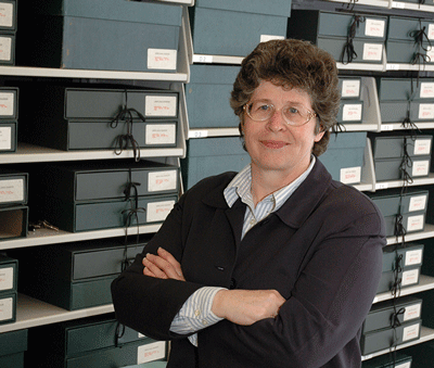 A woman, Susan Hodson, stands in front shelved folders. She's wearing glasses and a black sweater over a white shirt. She has her arms crossed in front of her