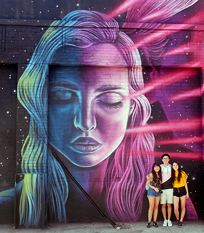 Three students pose for a photograph in front of a wall-sized mural of a woman in downtown Los Angeles.