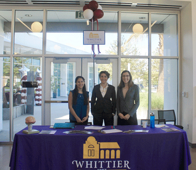 Three women stand behind a table with a sign that reads "Whittier College Established 1887" 
