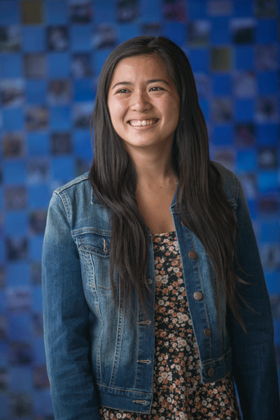 female student wearing a black dress with flowers and a denim jacket stands in front of a blurred, blue background