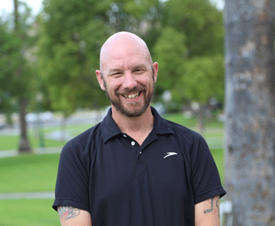 A man wearing a black polo shirt stands smiling at the camera with his arms at his side.