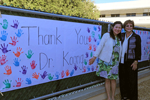 Two women stand next to each other. To the left a sign that reads "Thank you Mrs. Koong" hangs from the fence