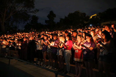 A group of students stand in an outdoor stadium. The sky is dark and they are holding candles. 