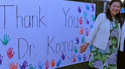 Two women stand next to each other. To the left of them, a sign that reads "Thank you Mrs. Koong" hangs on a fence