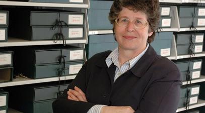 A woman, Susan Hodson, stands in front shelved folders. She's wearing glasses and a black sweater over a white shirt. She has her arms crossed in front of her