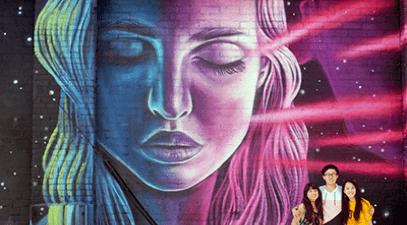 Three students pose for a photograph in front of a wall-sized mural of a woman in downtown Los Angeles.