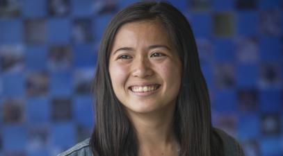 female student wearing a black dress with flowers and a denim jacket stands in front of a blurred, blue background