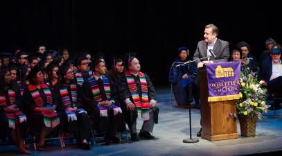 students in caps and gowns sit on stage listening to speaker at the podium