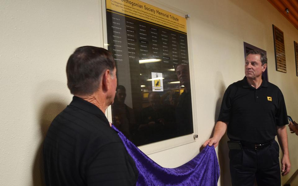 Two men hold a purple curtain in front of a plaque.